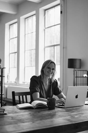 Vrouw achter haar laptop aan een werktafel, met een notitieboek naast zich